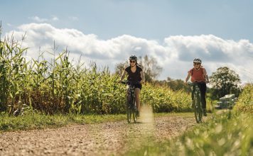 Zwei Frauen fahren mit dem Fahrrad einen Schotterweg hinunter. Links und rechts vom Weg wächst Mais.