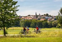 Eine Gruppe von vier Radfahrern fährt von rechts nach links im Vordergrund duch das Bild. Der gerade verlaufende Radweg geht durch grüne Wiesen und Felder. Am linken Bildrand steht ein großer, grüner Laubbaum, am rechten ein kleiner. Im Hintergrund steigt nach einer Baumreihe die kleine Stadt Herbstein auf einem Bergrücken emport. Im Zentrum sind deutlich zwei Kirchtürme und ein Turm der Stadtmauer zu erkennen.