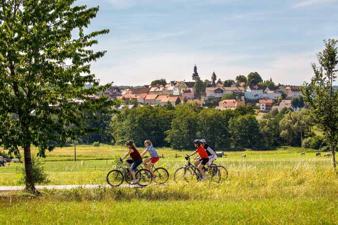 Auf dem Vulkan-Radweg vor Herbstein Eine Gruppe von vier Radfahrern fährt von rechts nach links im Vordergrund duch das Bild. Der gerade verlaufende Radweg geht durch grüne Wiesen und Felder. Am linken Bildrand steht ein großer, grüner Laubbaum, am rechten ein kleiner. Im Hintergrund steigt nach einer Baumreihe die kleine Stadt Herbstein auf einem Bergrücken emport. Im Zentrum sind deutlich zwei Kirchtürme und ein Turm der Stadtmauer zu erkennen.
